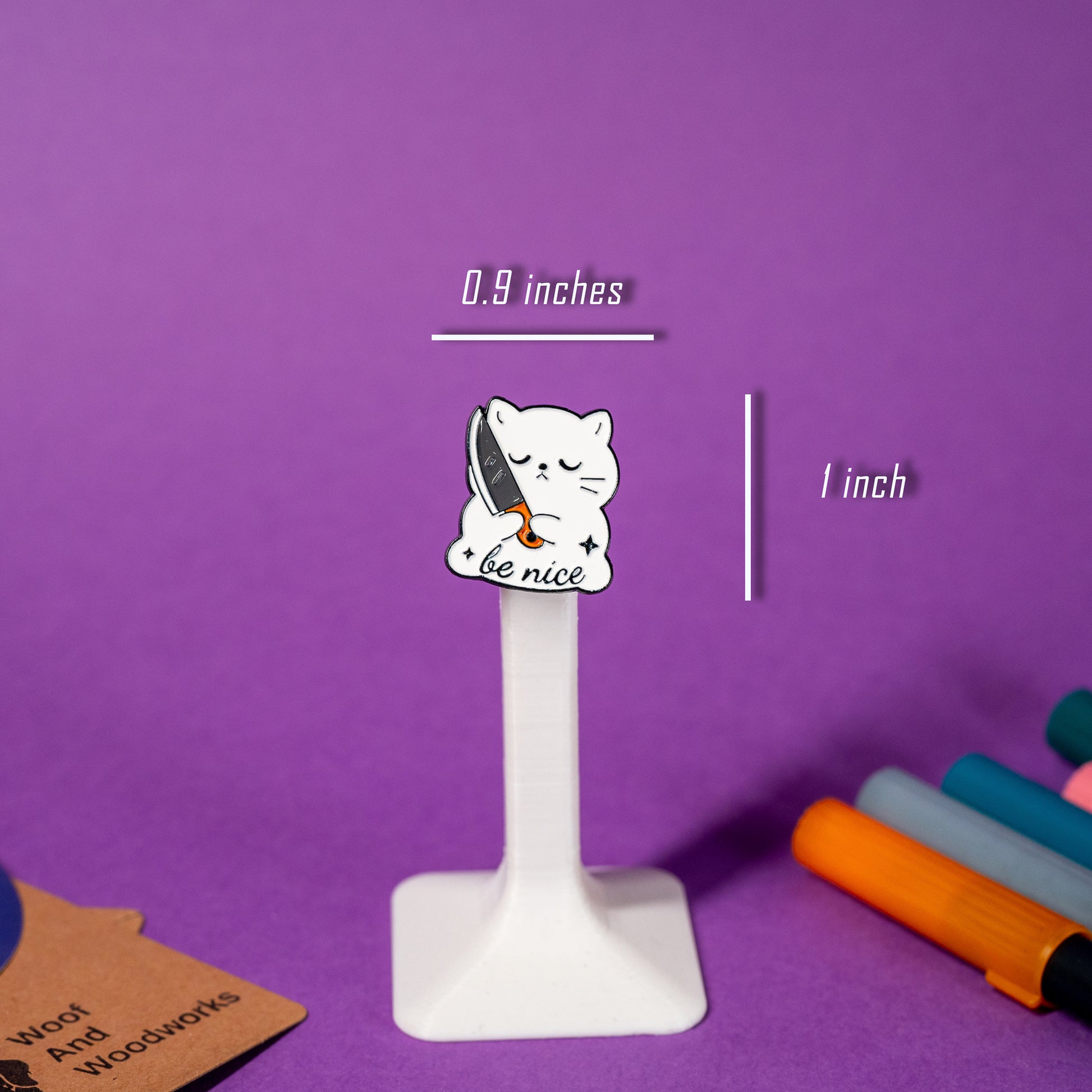 Enamel pin of a white cat hugging a knife with a neutral expression and the phrase Be Nice.