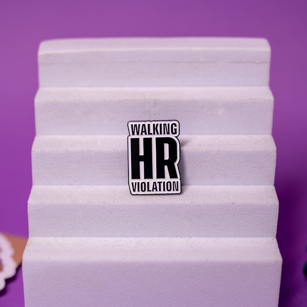 Walking HR Violation enamel pin displayed on foam stand, simple black and white text design.