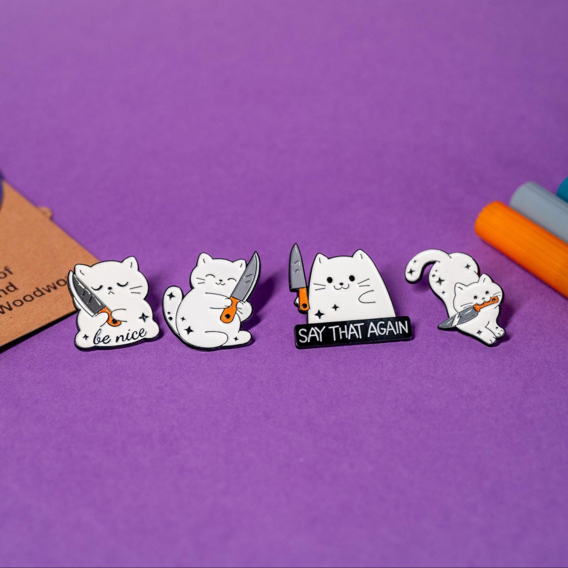 Set of 4 white cat enamel pins featuring playful designs with cats and knives, including 'Say That Again,' 'Be Nice,' hugging knife, and jumping cat.