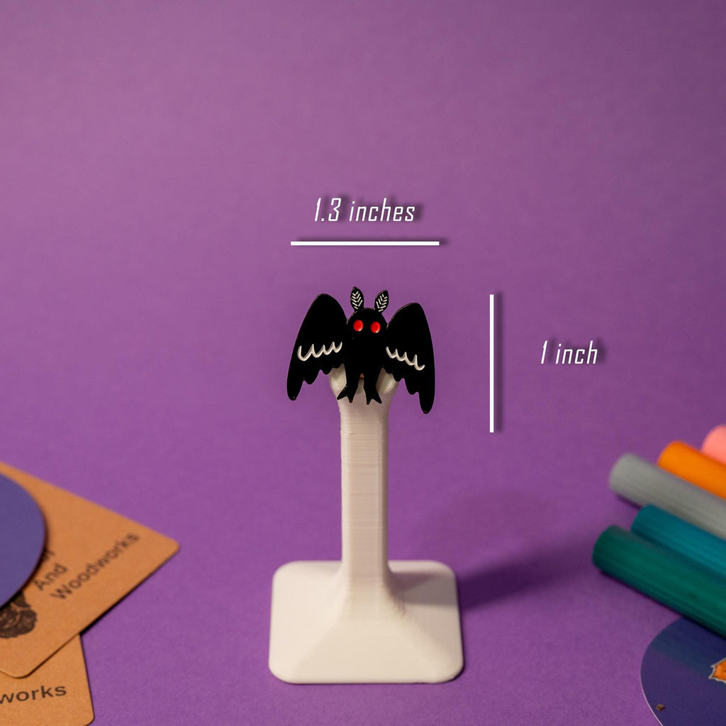 Mothman enamel pin displayed on a stand, highlighting the cryptid creature’s iconic silhouette.
