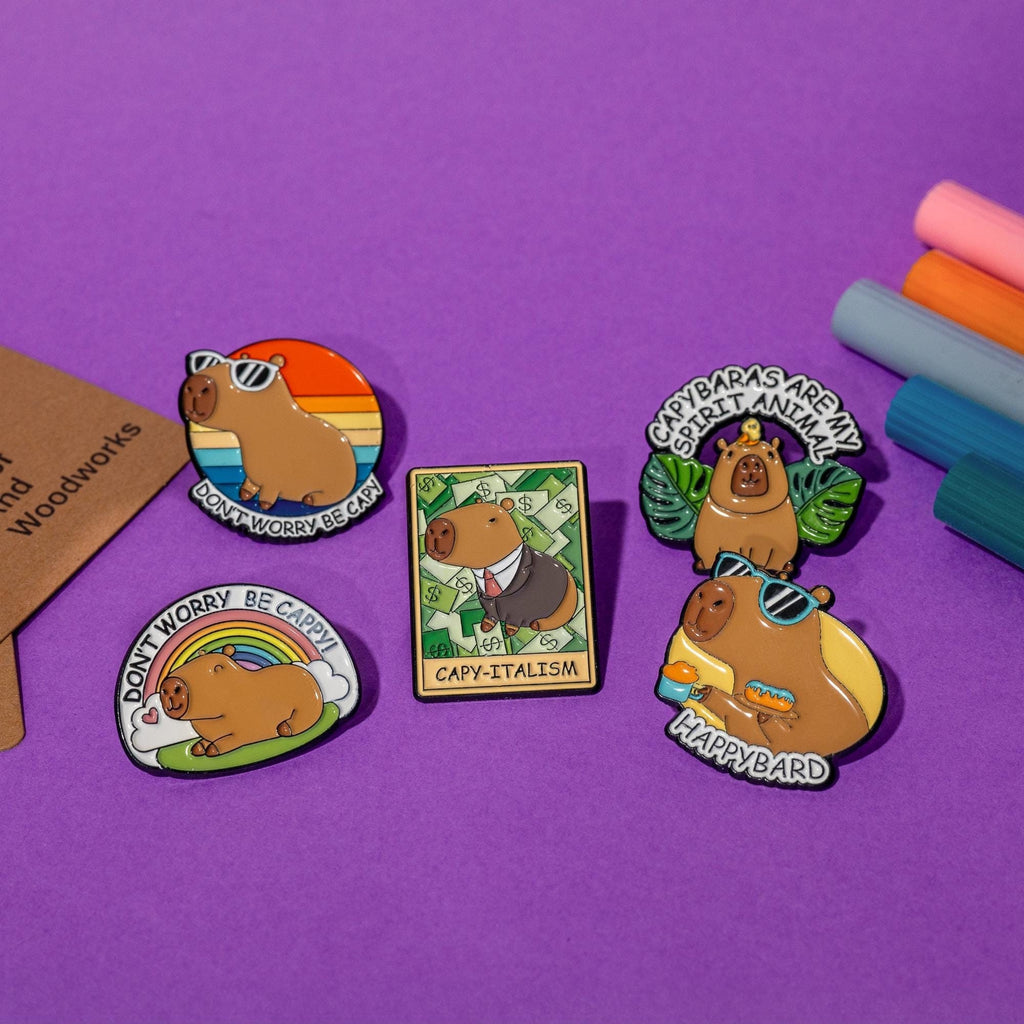Set of 5 enamel pins featuring funny capybara designs: Don’t Worry Be Capy, Capy-Italism, Happybard, Capybaras Are My Animal Spirit, and Don’t Worry Be Capppy!