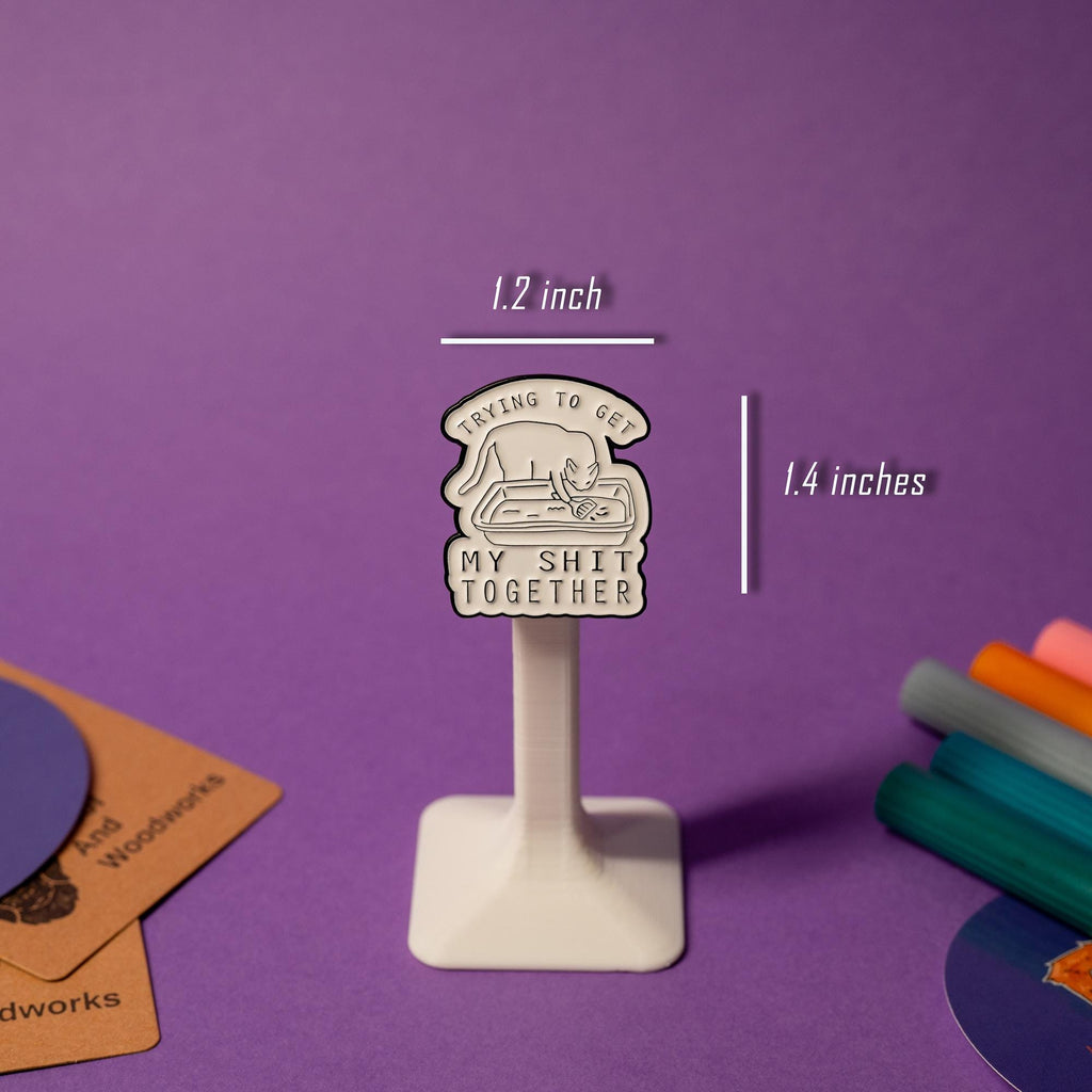 Close-up of enamel pin with a quirky cat scooping litter, showcasing the text Trying to get my sh*t together.