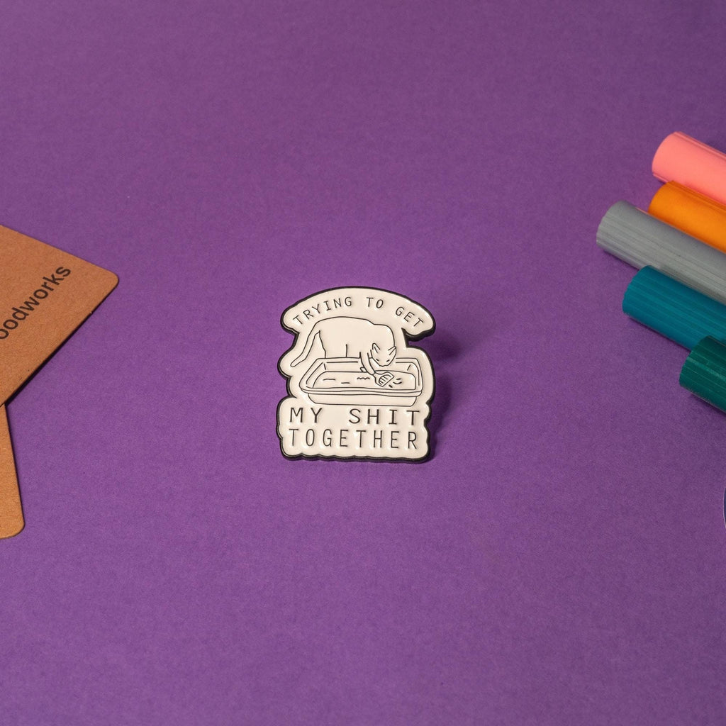 Funny enamel pin of a cat in a litter box with the phrase Trying to get my sh*t together, perfect for humor fans.