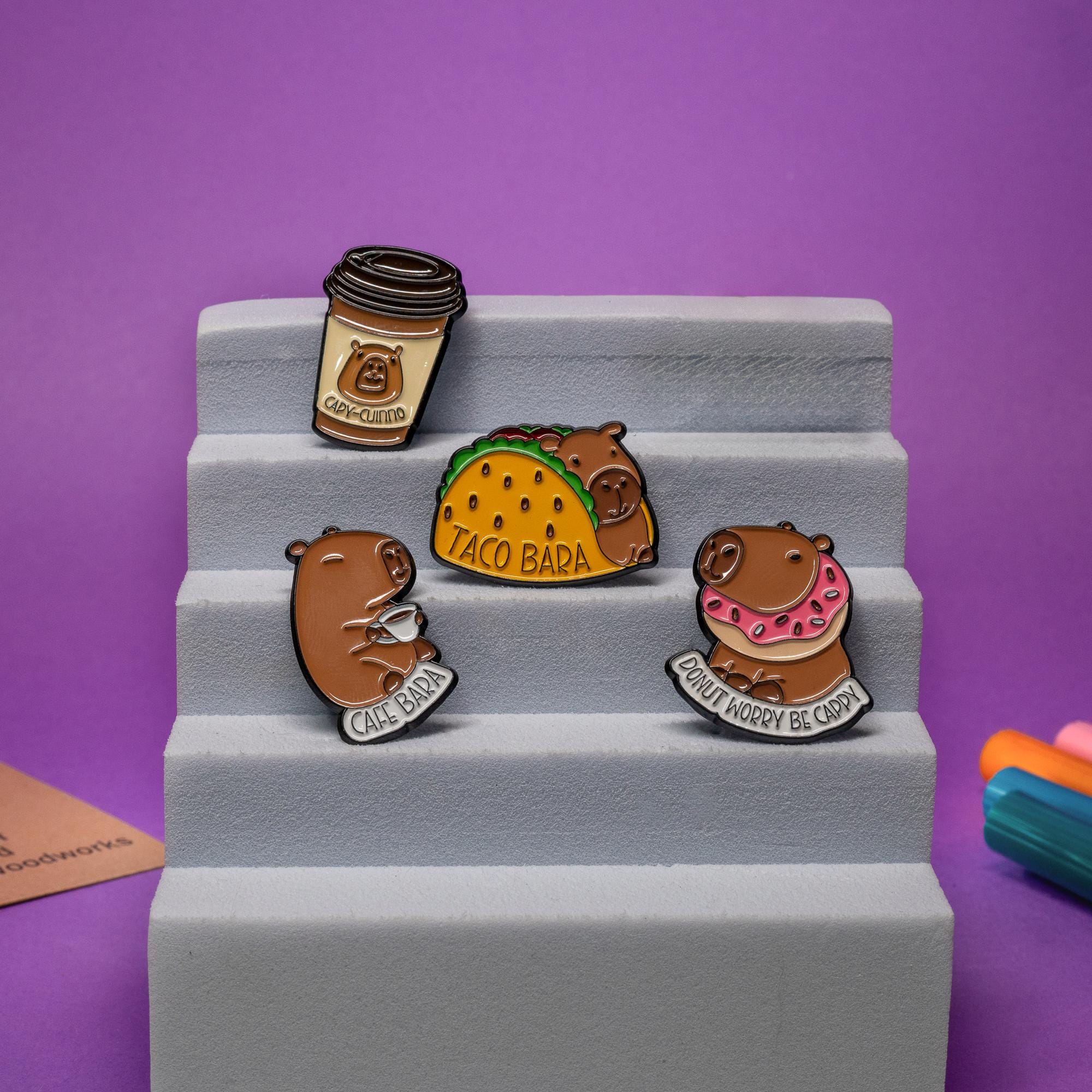 Capybara enamel pins displayed together, including Taco Bara and Donut Worry Be Cappy designs.