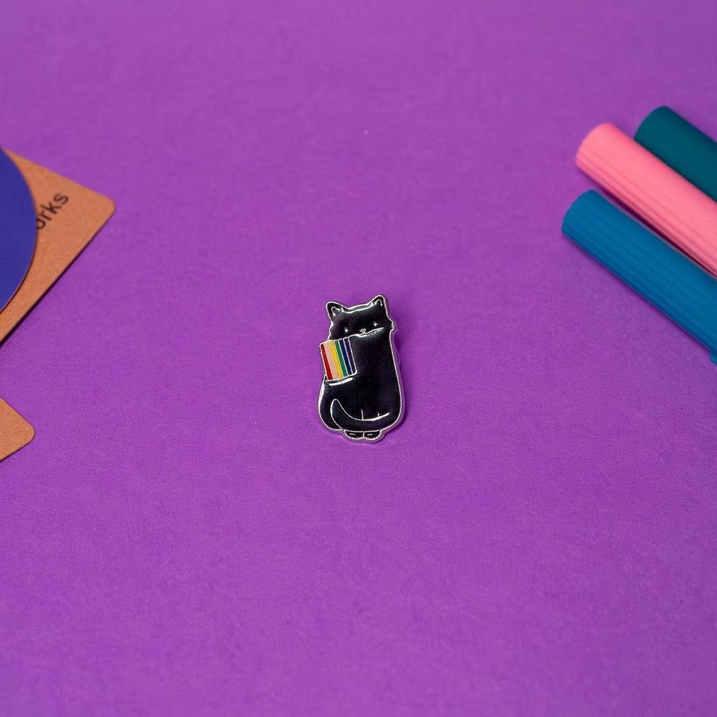 LGBTQ Pride enamel pin featuring a black cat sitting and holding a rainbow flag, displayed on a flat surface to highlight its vibrant design.