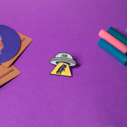 Alien cat enamel pin featuring a green alien-like cat in a UFO abducting a black Earth cat with a yellow beam, displayed on a flat surface to highlight its quirky design.