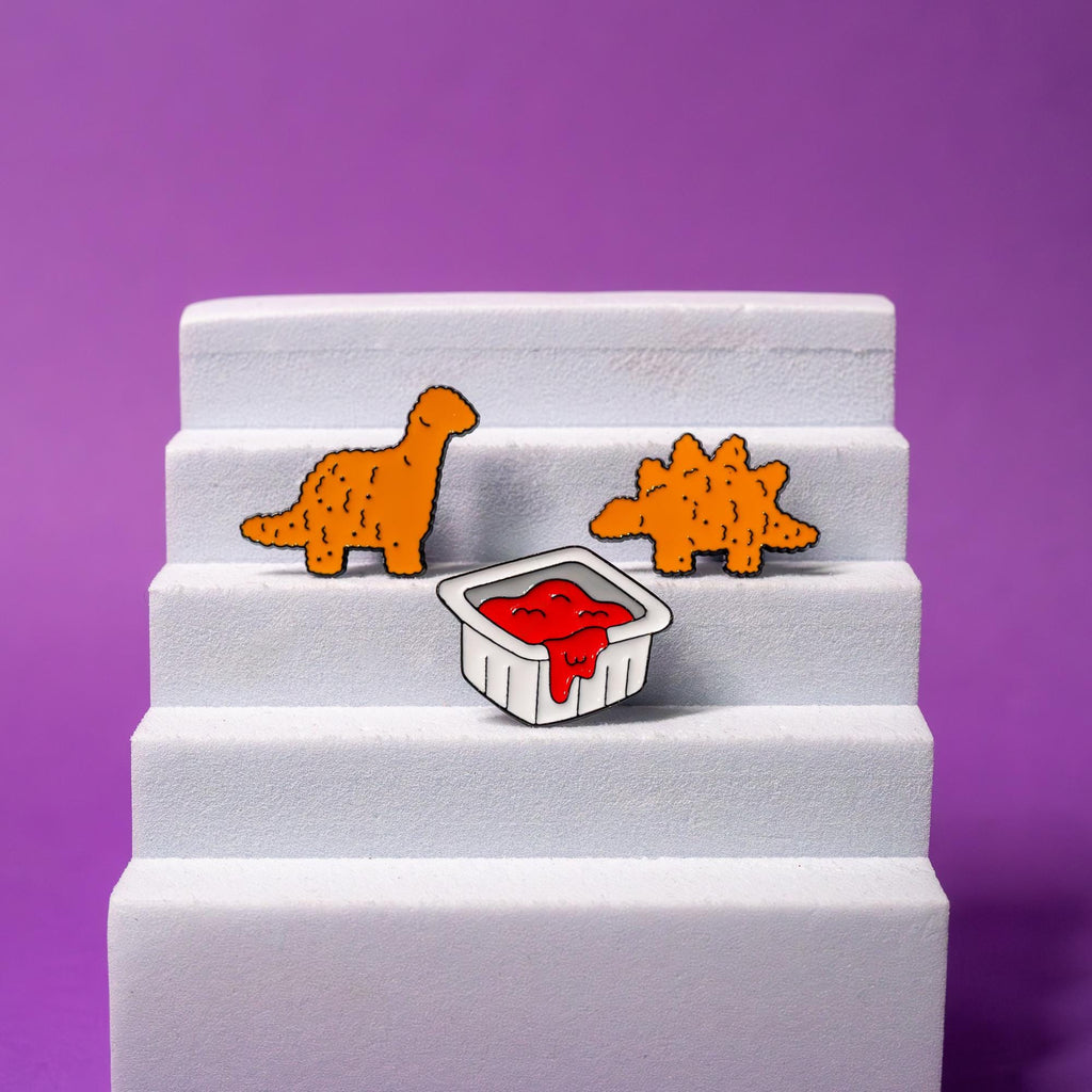 Dino Nugget enamel pin set displayed on a foam stand, featuring the stegosaurus, brachiosaurus, and ketchup packet pins in a playful arrangement.