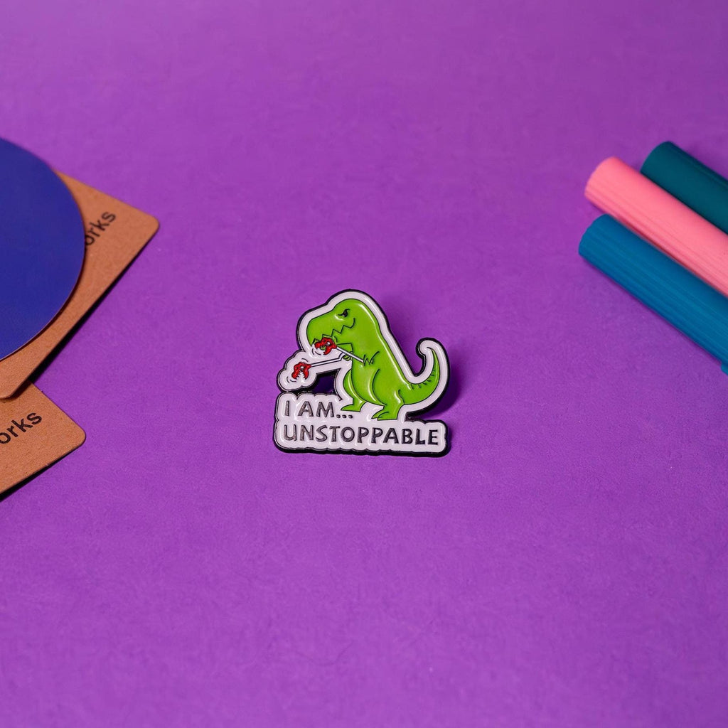 Enamel pin featuring a cute T-Rex with extended hand grabbers and the text &#39;I AM... UNSTOPPABLE,&#39; displayed on a flat surface to highlight its quirky design.