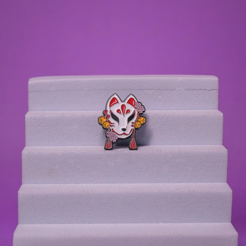 Traditional Japanese Kitsune mask enamel pin displayed on a foam stand, emphasizing its bold design and cultural significance.