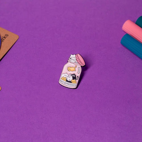 Enamel pin featuring a bottle labeled &#39;Anti Depressants&#39; with an unscrewed lid and kawaii-style cats inside, displayed on a flat surface to highlight its adorable design.