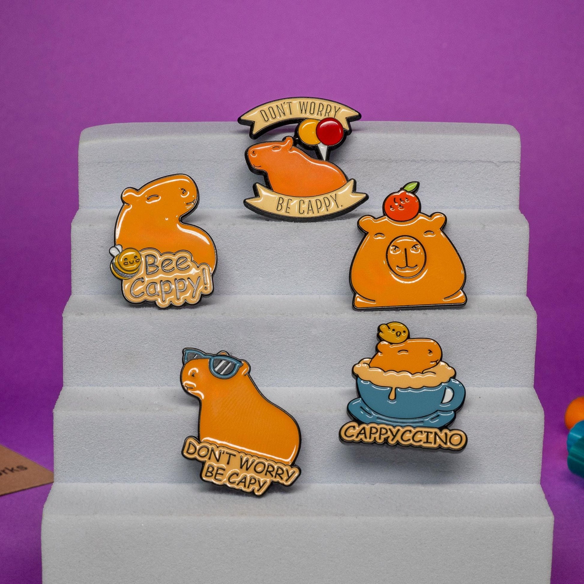 Friendly Capybara enamel pin set displayed on a foam stand, showcasing all five designs with vibrant colors and cute capybara themes.