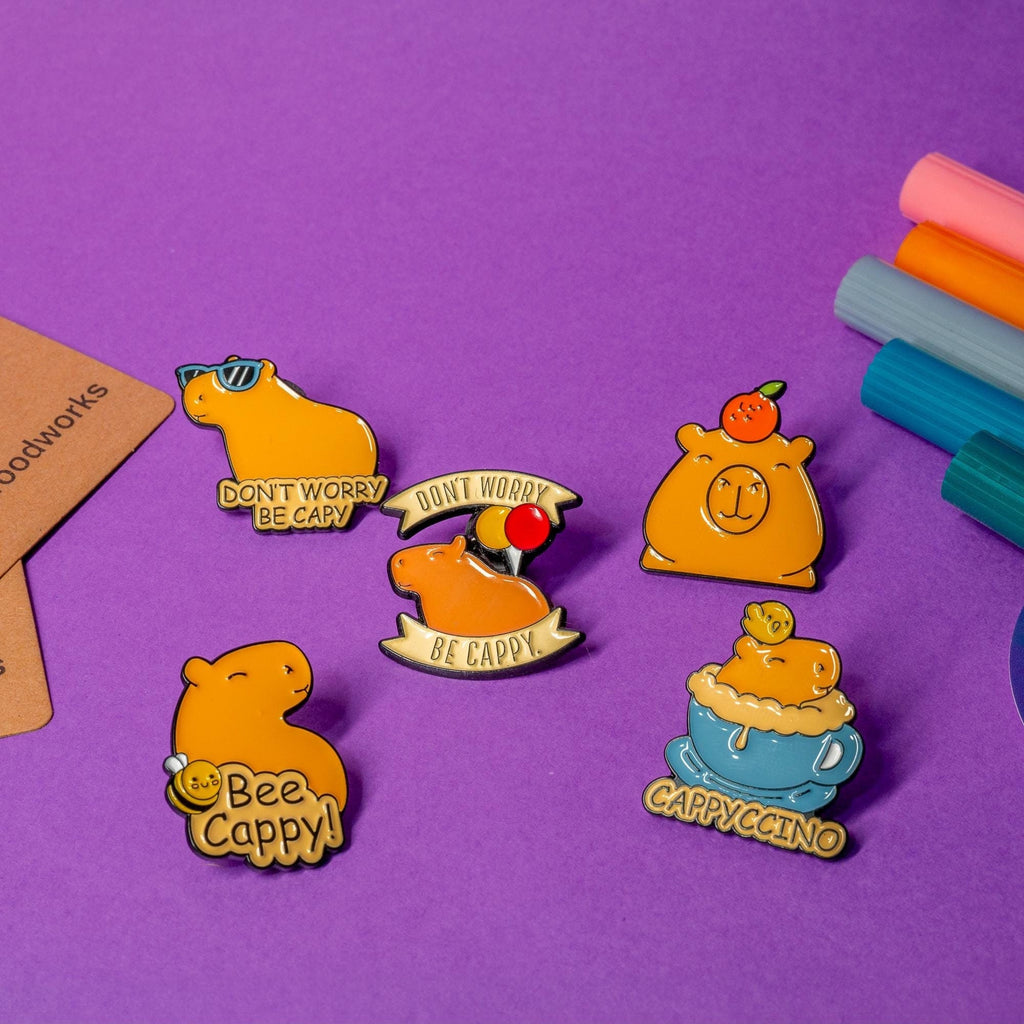 Set of 5 Friendly Capybara enamel pins displayed on a flat surface, featuring quirky designs including No Text Capybara, Bee Cappy!, Cappyccino, and Dont Worry variations.