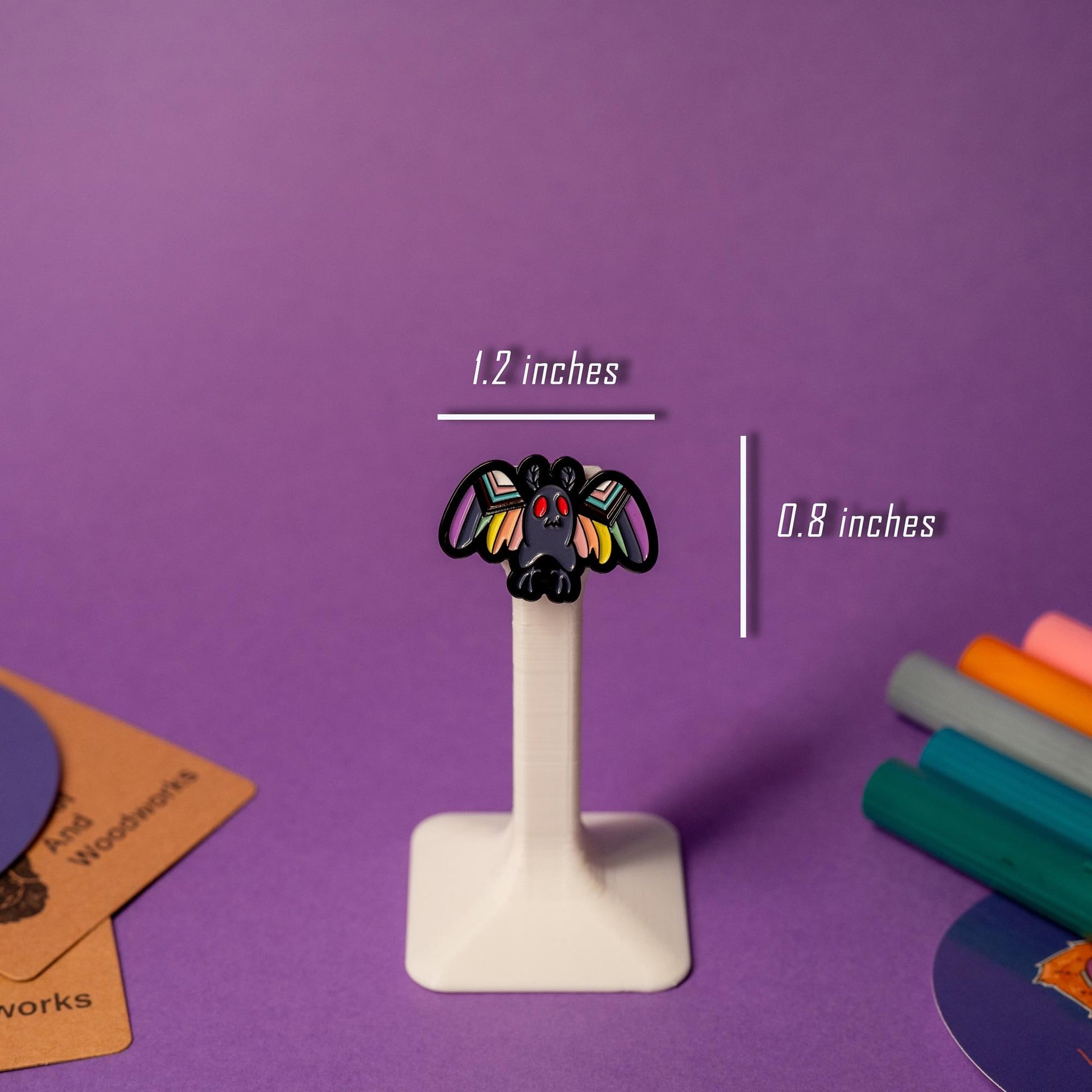 Mothman enamel pin with LGBTQ+ flag design displayed on a stand, celebrating inclusivity and mystery.