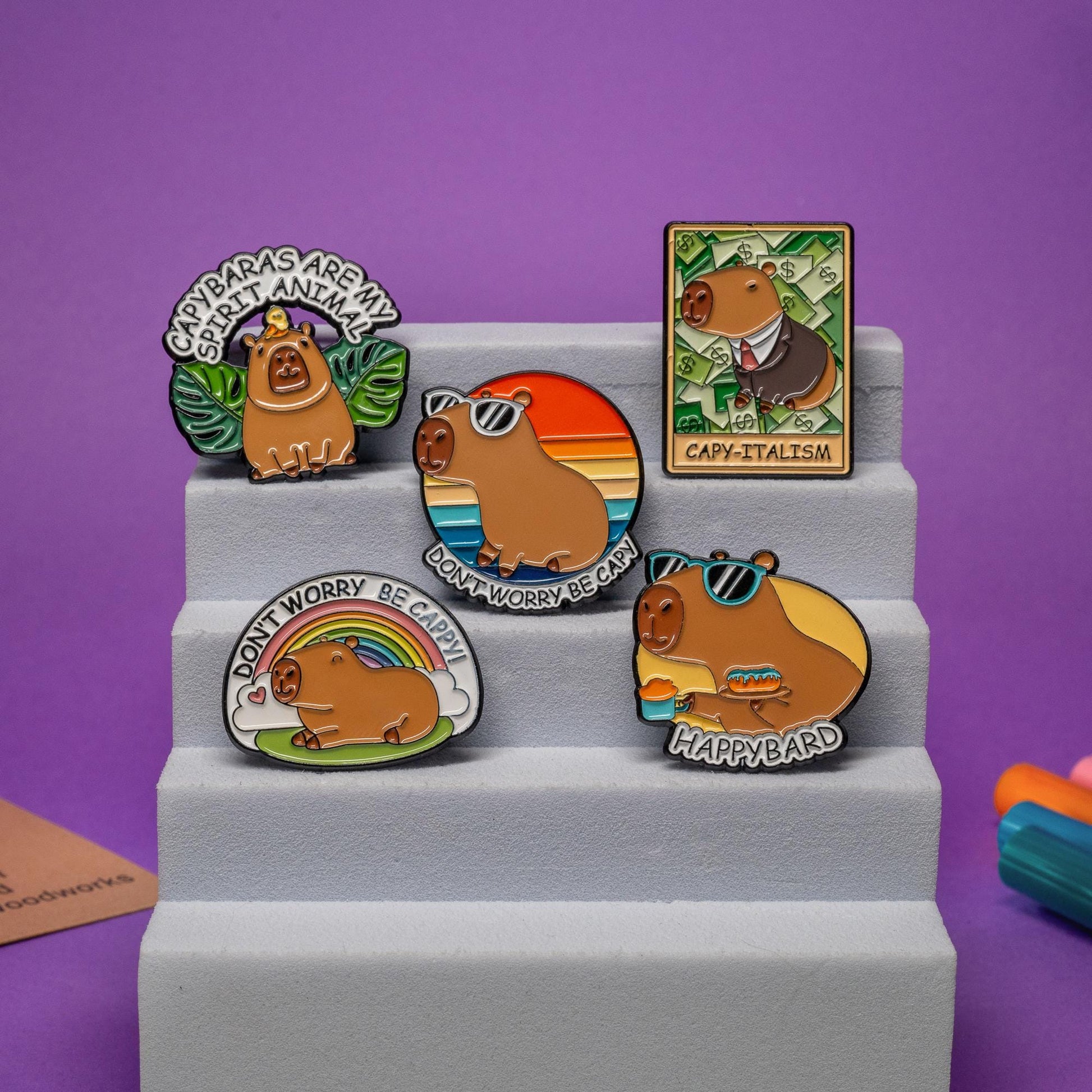 Group photo of 5 capybara enamel pins, including sunglasses, suit, coffee and donut, rainbow, and animal spirit themes.
