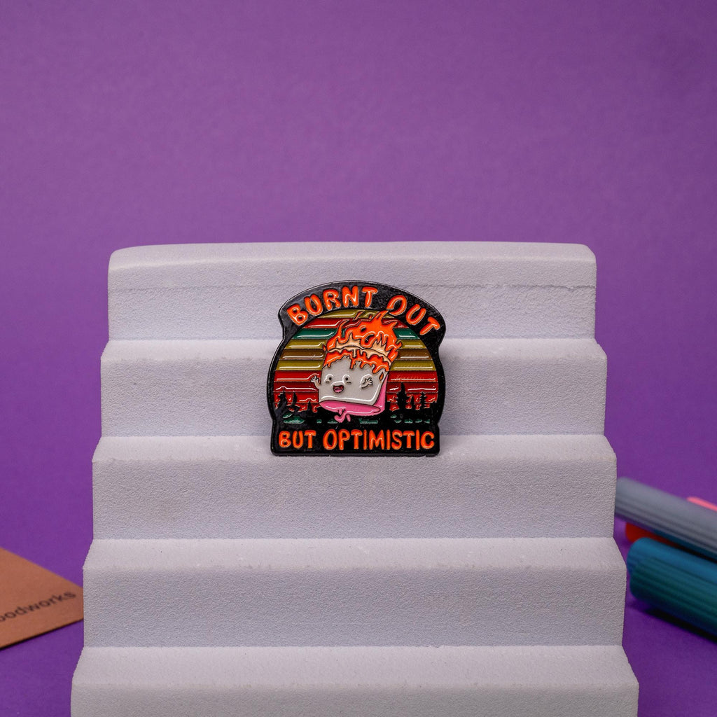 Close-up of a marshmallow enamel pin on fire, showcasing bold colors and the text Burnt Out but Optimistic.