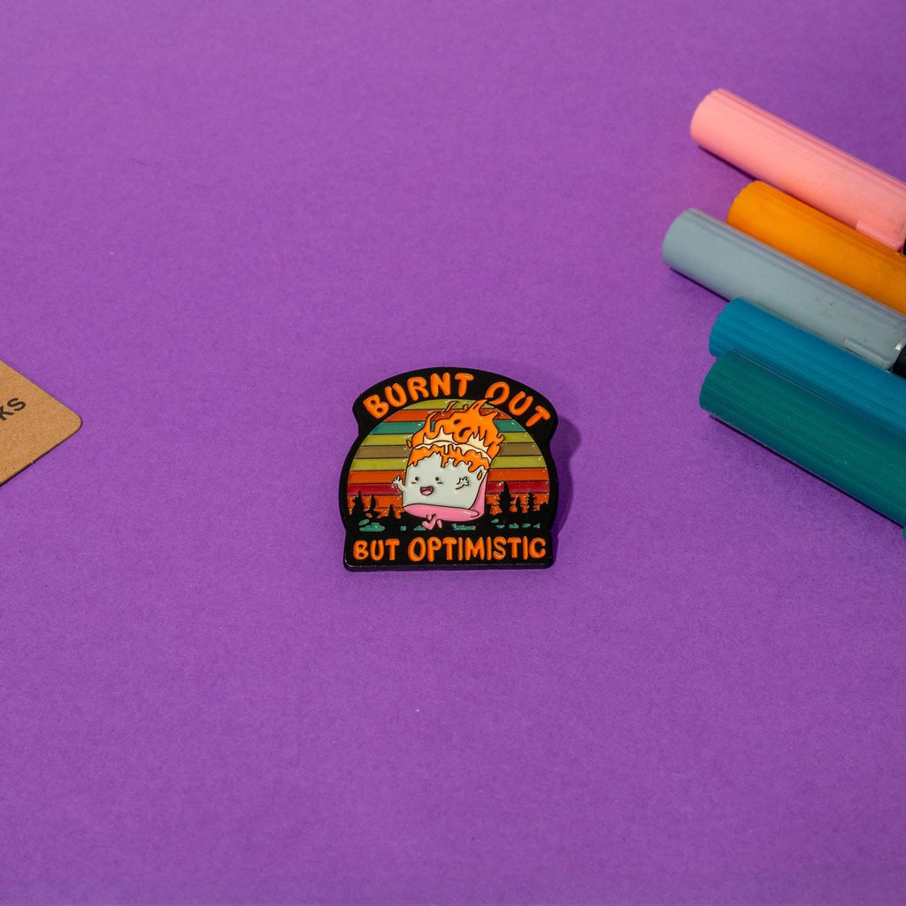 Funny marshmallow enamel pin with flames and the phrase Burnt Out but Optimistic, perfect for humor and camping fans.