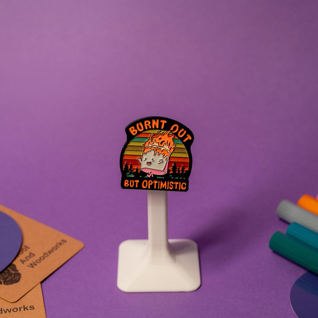 Enamel pin featuring a marshmallow running on fire with the humorous text Burnt Out but Optimistic.