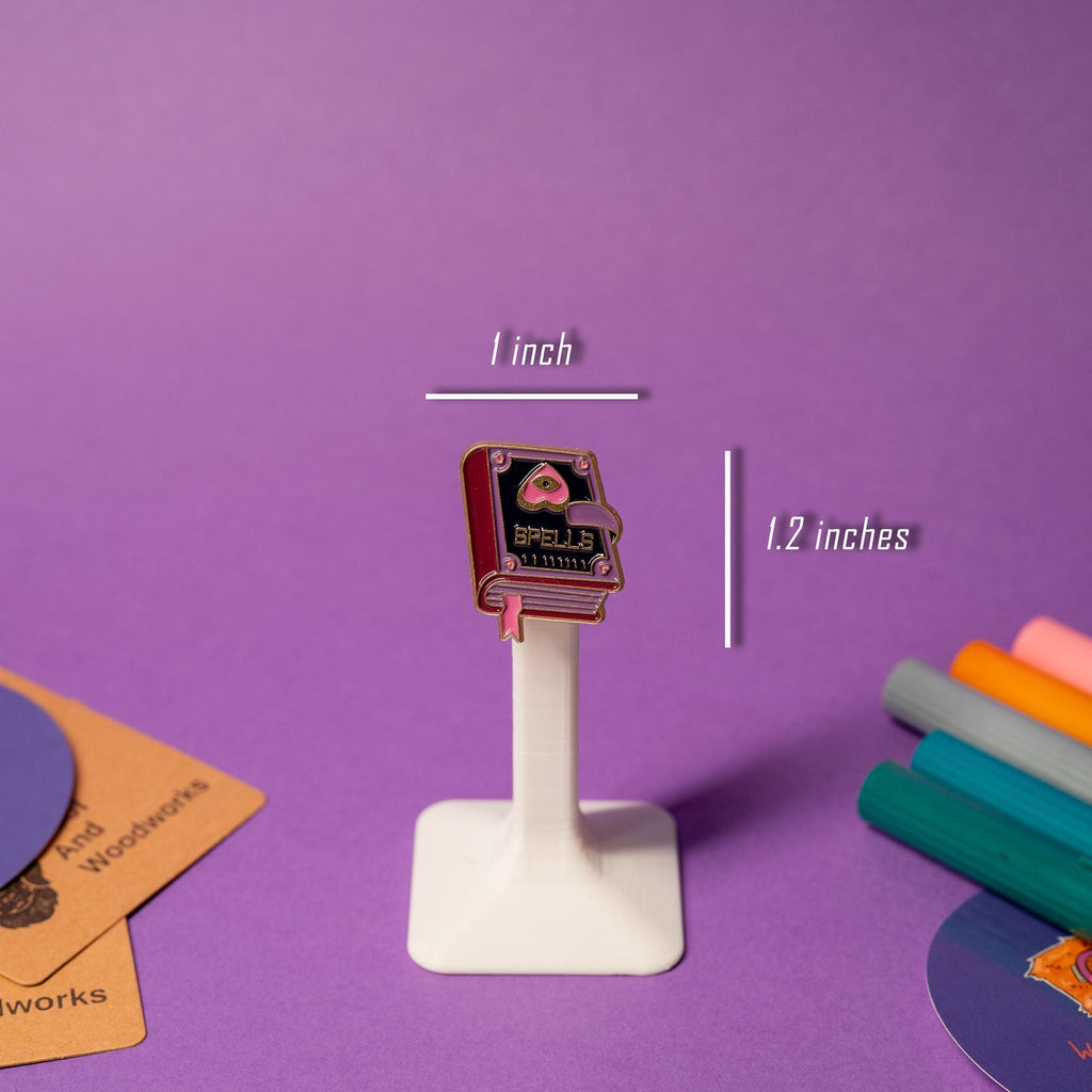 Enamel pin of a spell book with the word Spells, designed in a witchy purple-pink color scheme.