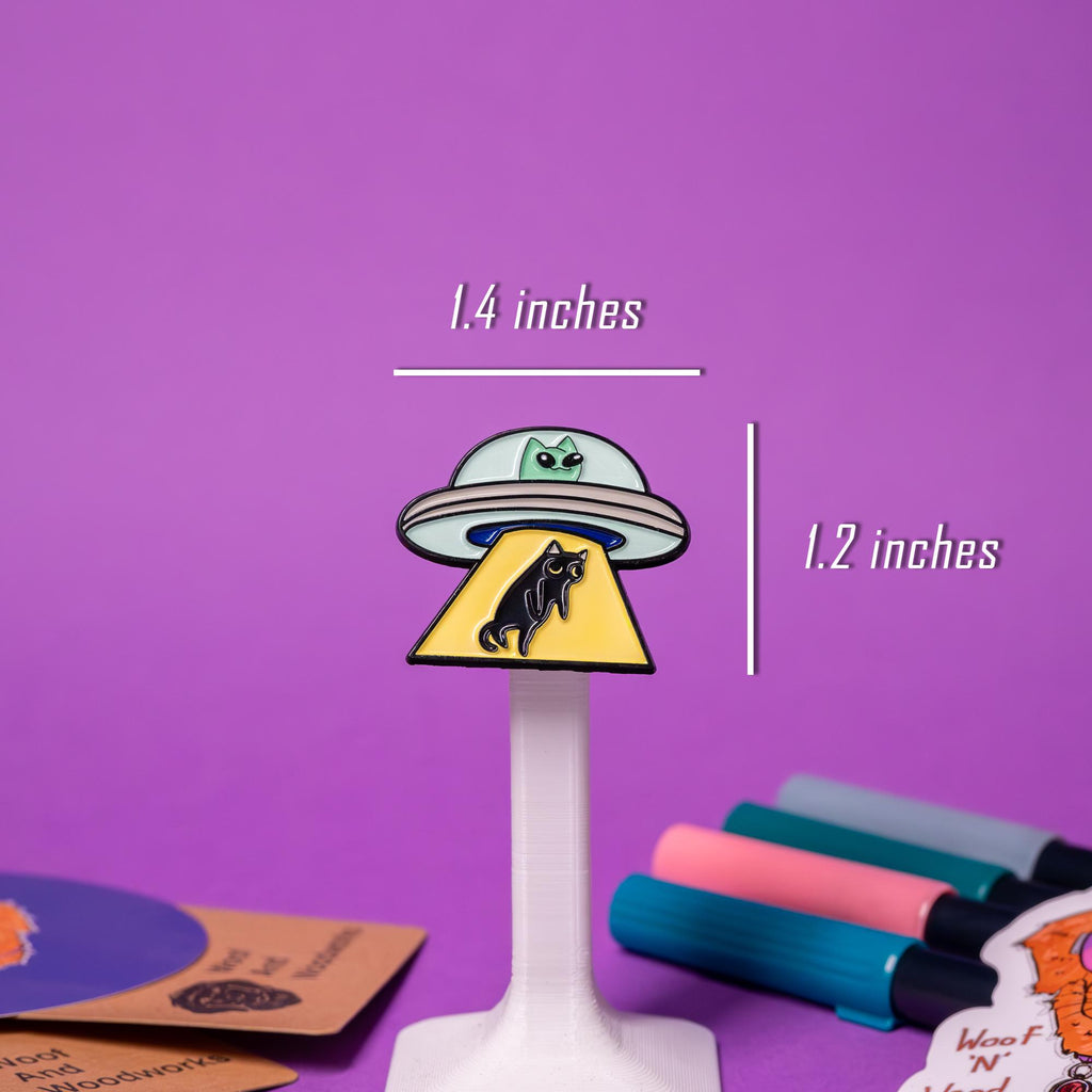 Close-up of the alien cat enamel pin, showcasing intricate details of the green cat, flying saucer, and yellow abduction beam.