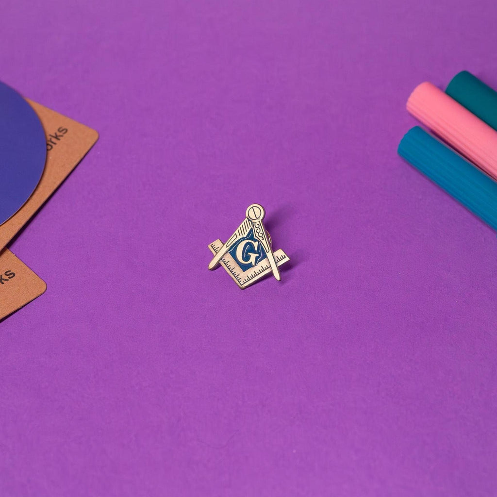 Freemason enamel pin featuring the Square & Compass symbol with the letter G in the center, displayed on a flat surface to showcase its elegant design.