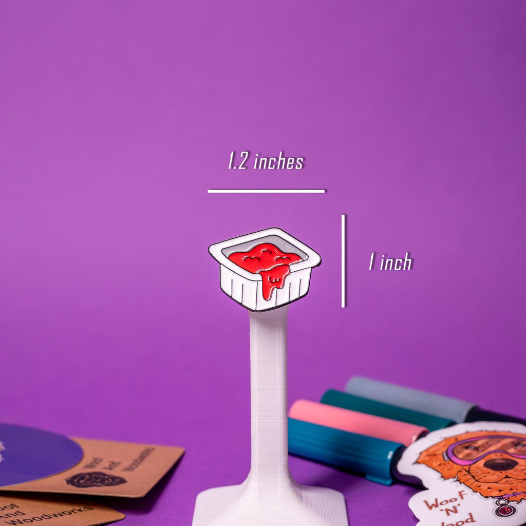 Close-up of the fast-food-style ketchup packet enamel pin, featuring a bright red design and peel-back detail.