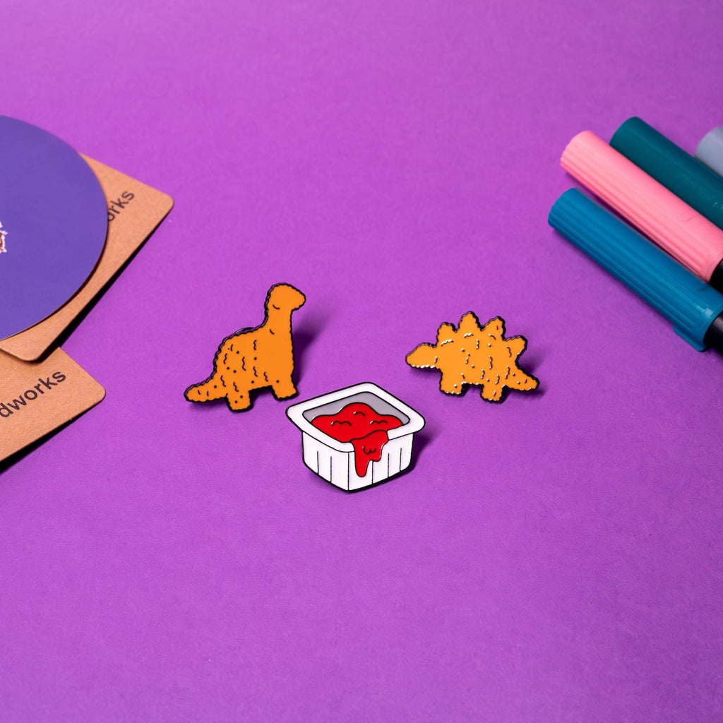Dino Nugget enamel pin set displayed on a flat surface, featuring a stegosaurus, a brachiosaurus, and a ketchup packet in vibrant detail.