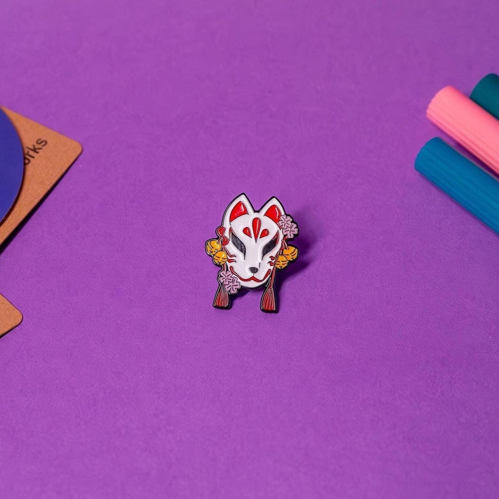 Enamel pin featuring a traditional Japanese Kitsune mask design, displayed on a flat surface to highlight its intricate details and elegant craftsmanship.